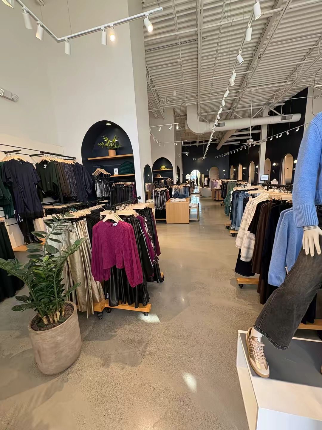 Interior of a retail clothing store featuring racks of apparel, a potted plant, and a mannequin in the foreground.