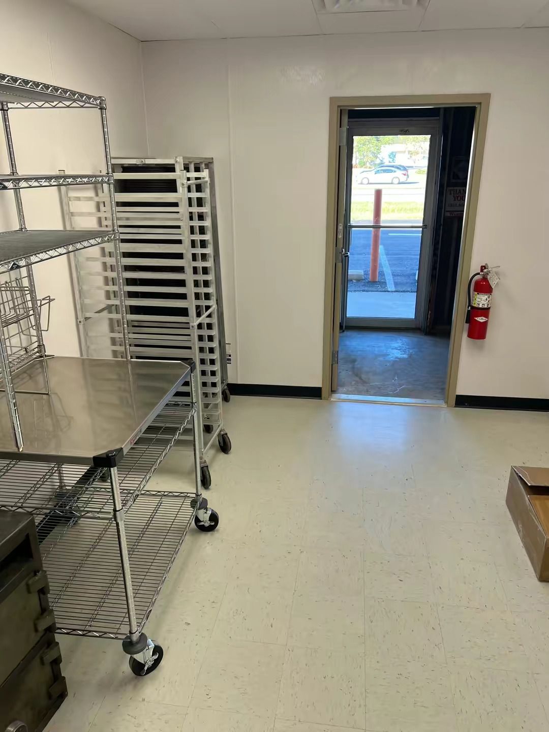 A commercial kitchen space with metal shelving, a rolling rack of baking trays, a doorway, and a red fire extinguisher.