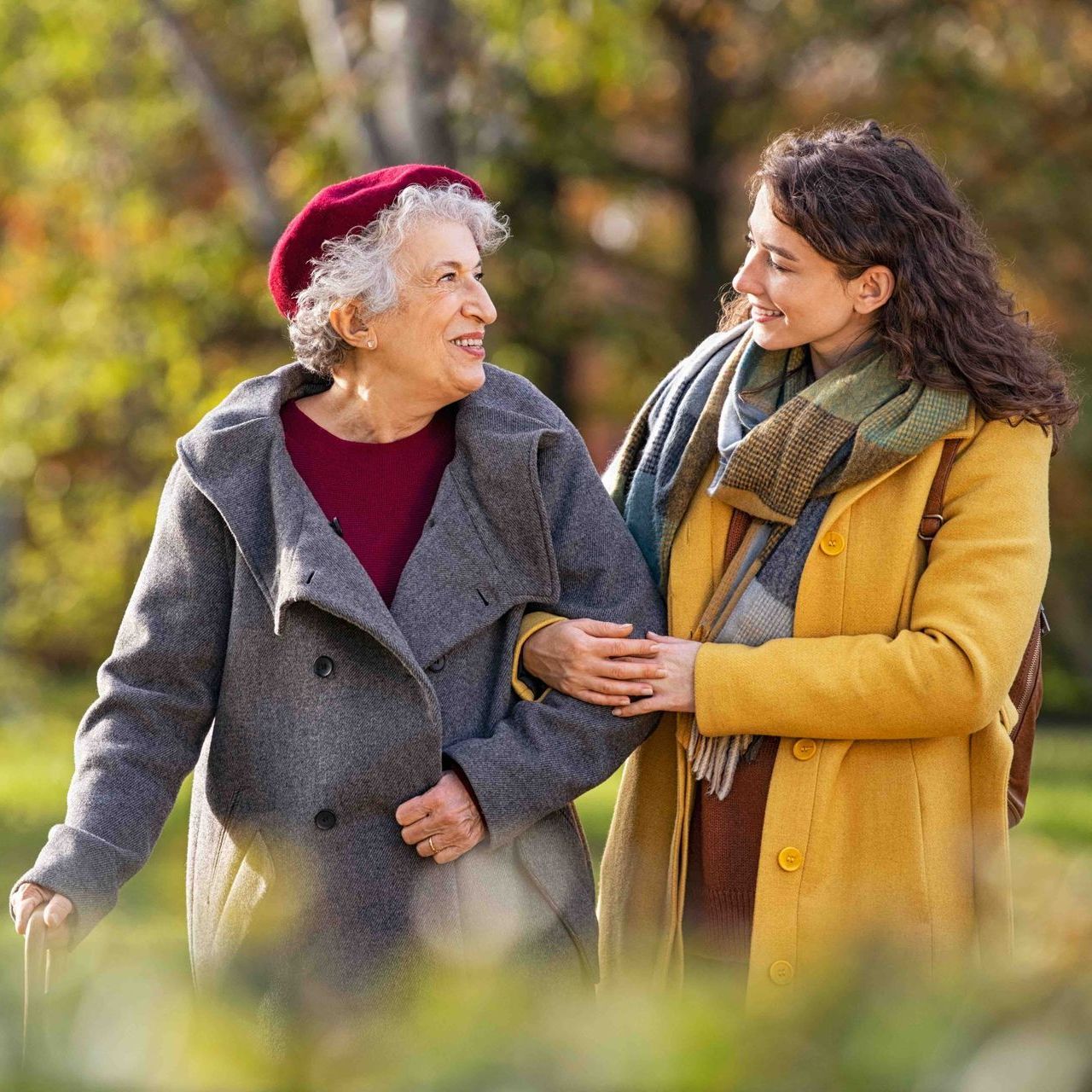 Woman helps senior walk outdoors, holding her arm. They smile, in a park.
