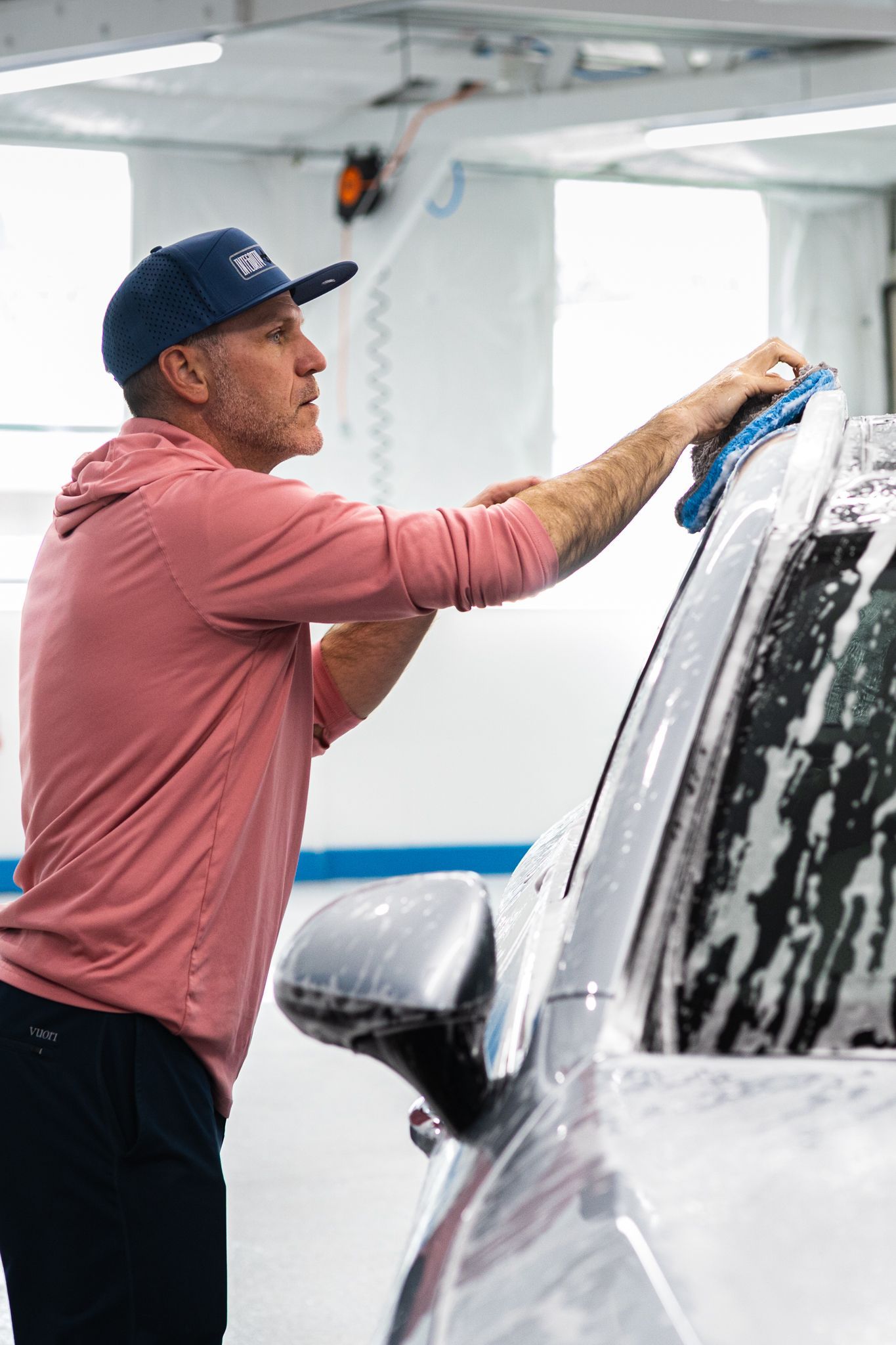 Man washing a car covered in soap at a carwash. He wears a cap and red shirt.