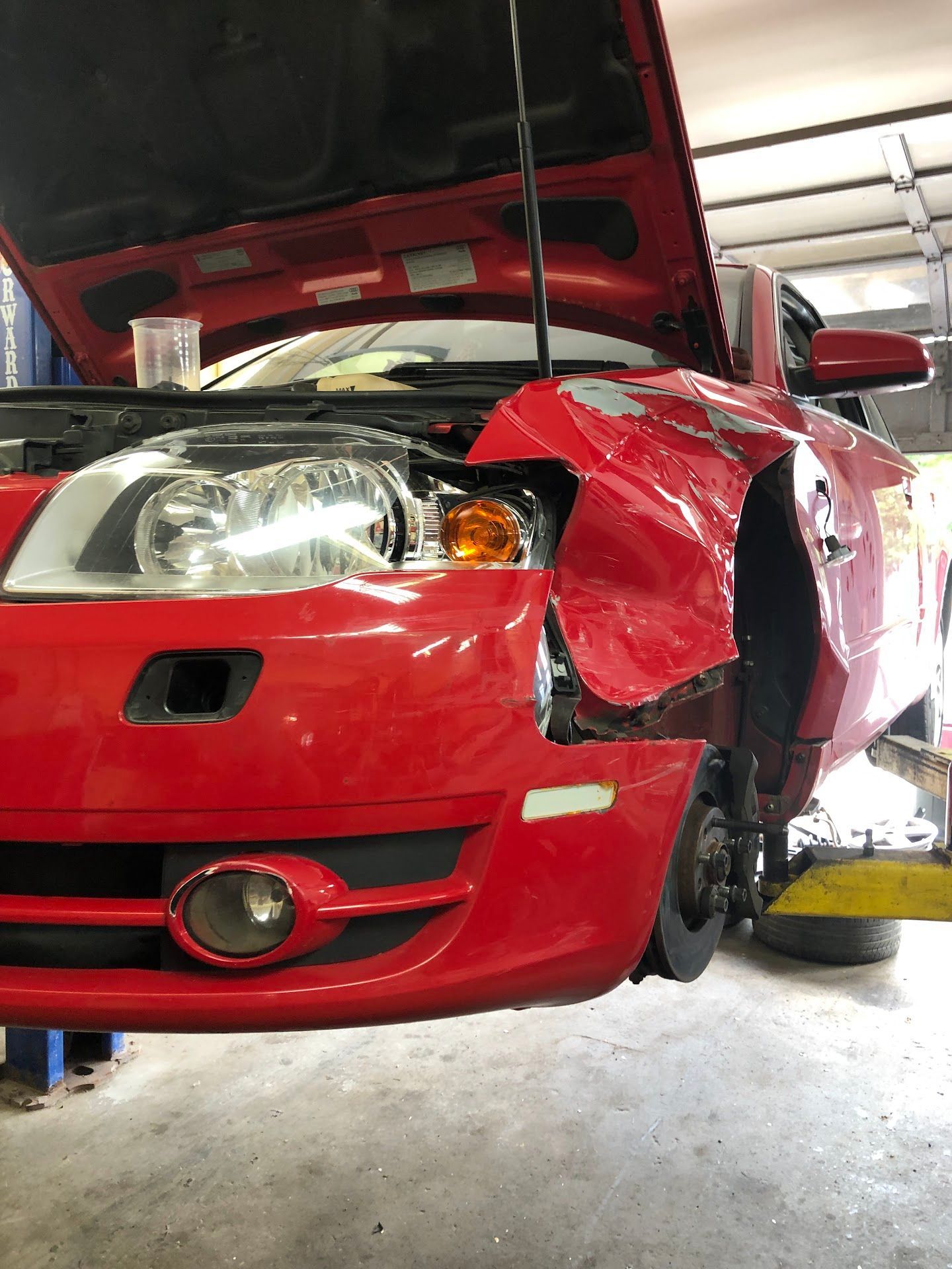 Red car with front-end damage is raised in a garage. Hood open, missing wheel, fender crumpled. | Rose Brothers Garage