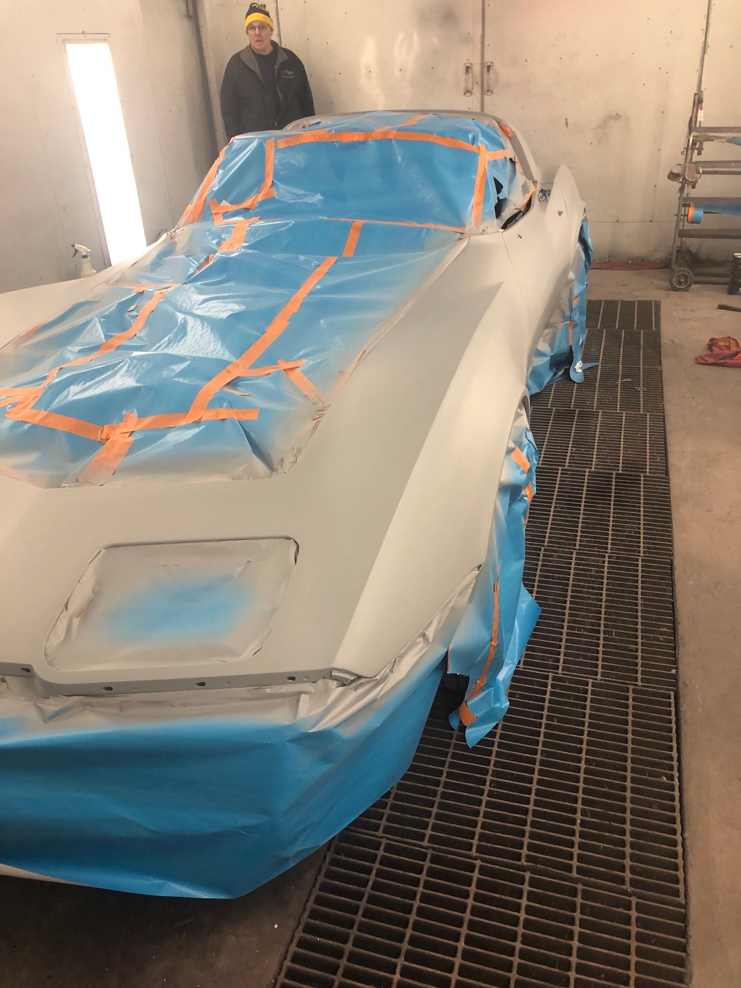 A Corvette is masked for painting inside a spray booth, with a worker in the background. | Rose Brothers Garage