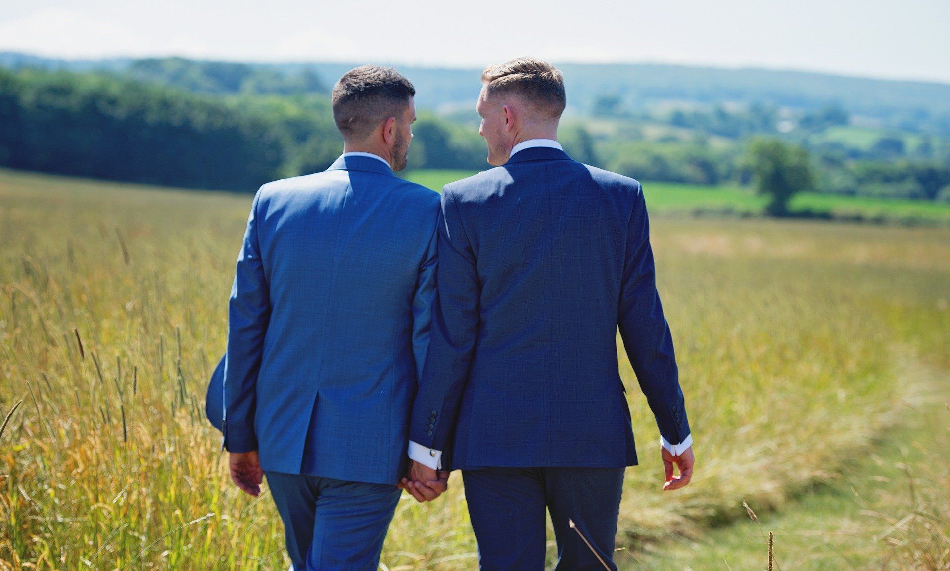 Two men in suits are walking through a field holding hands.