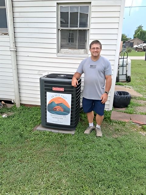 Man standing next to new air conditioning unit in front of a white building. Green grass.