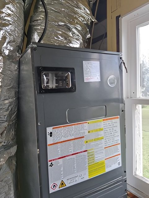 Gray furnace unit next to a window, with ductwork and warning labels visible.