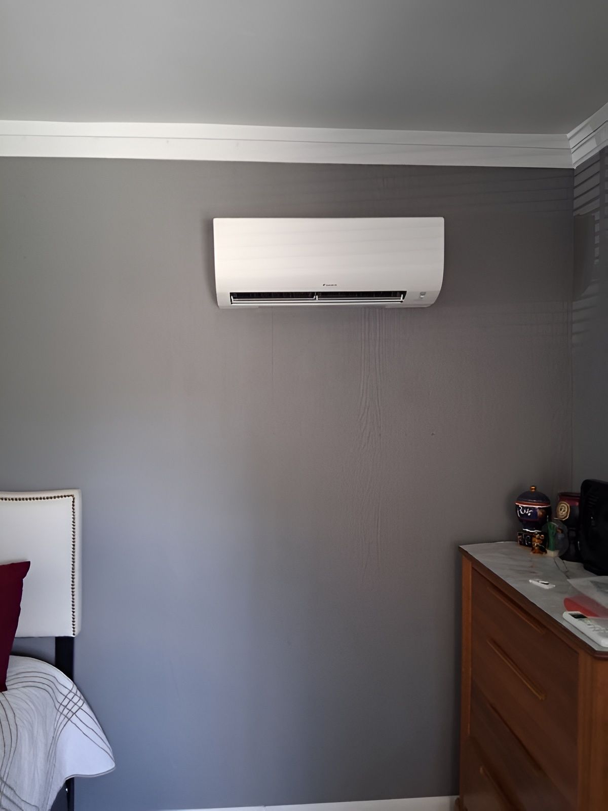 White air conditioner on a gray bedroom wall above a dresser and bed.