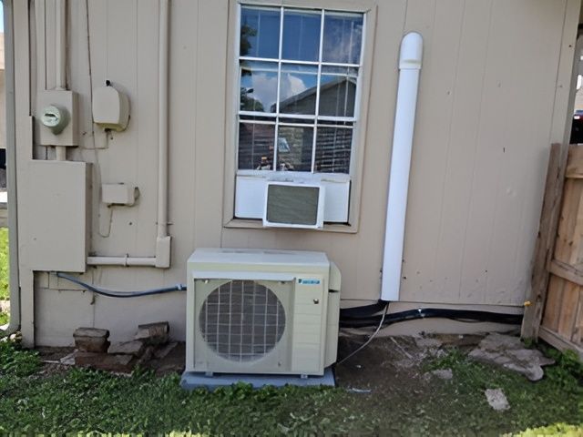 Outdoor AC unit below window with a window AC unit. White wall with electrical boxes and a vent pipe.