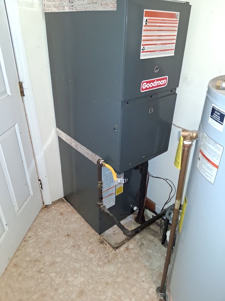 A Goodman furnace and water heater in a utility room. Gray appliances, white door, tan floor.