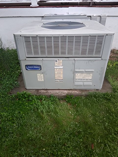 Gray air conditioning unit on a concrete base, set in a grassy yard, with a building in the background.