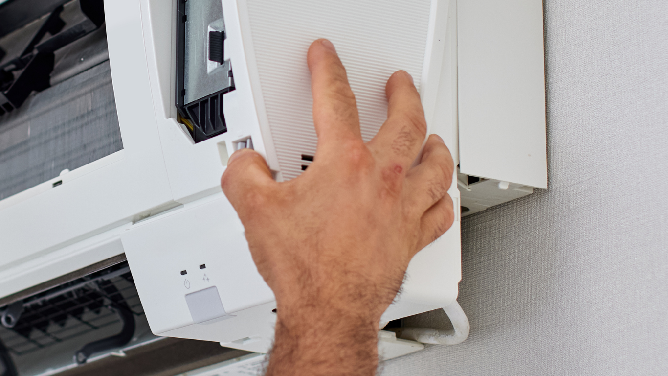 Hand opening the front panel of a white air conditioning unit on a white wall.