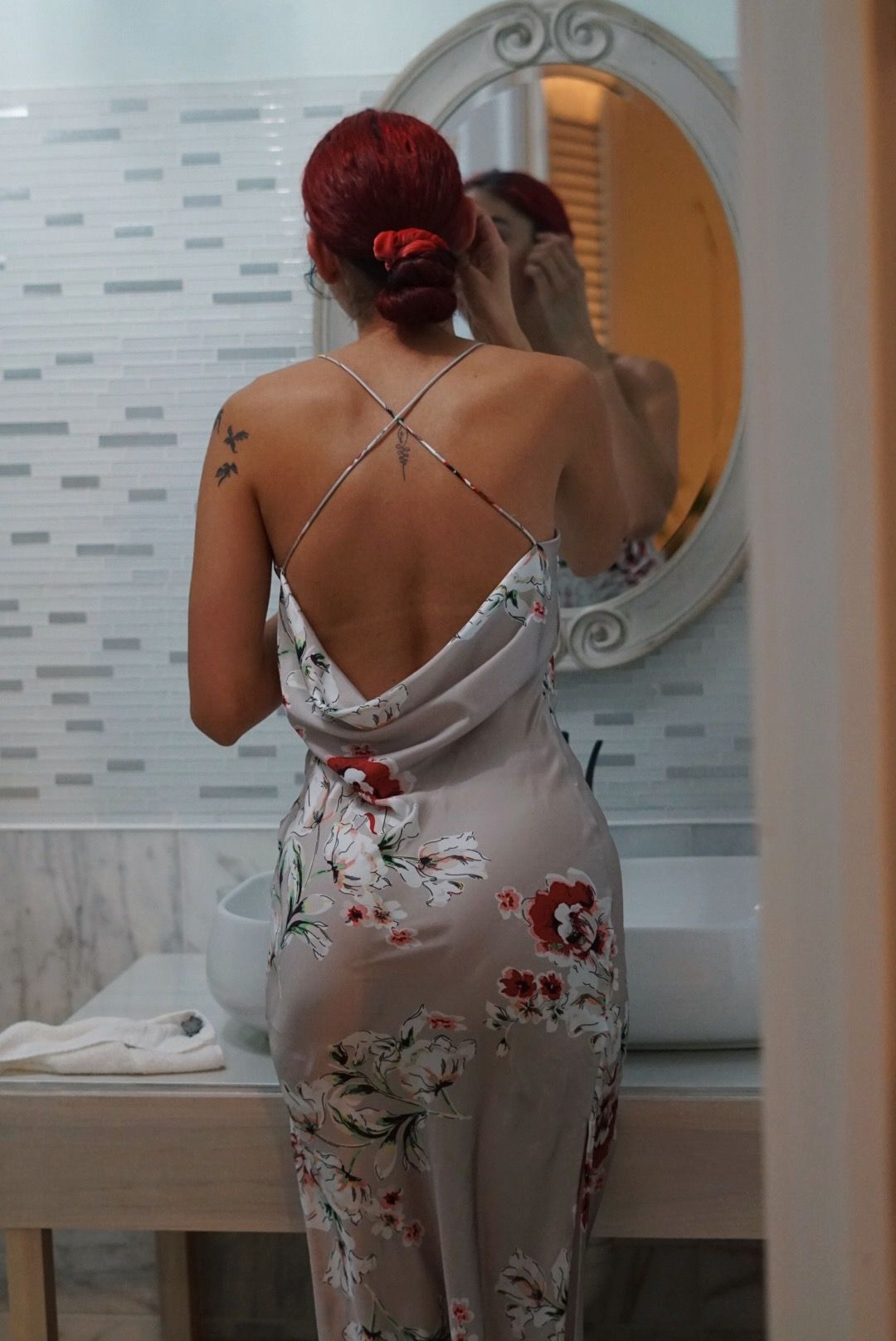 A woman in a floral dress is standing in front of a mirror in a bathroom.