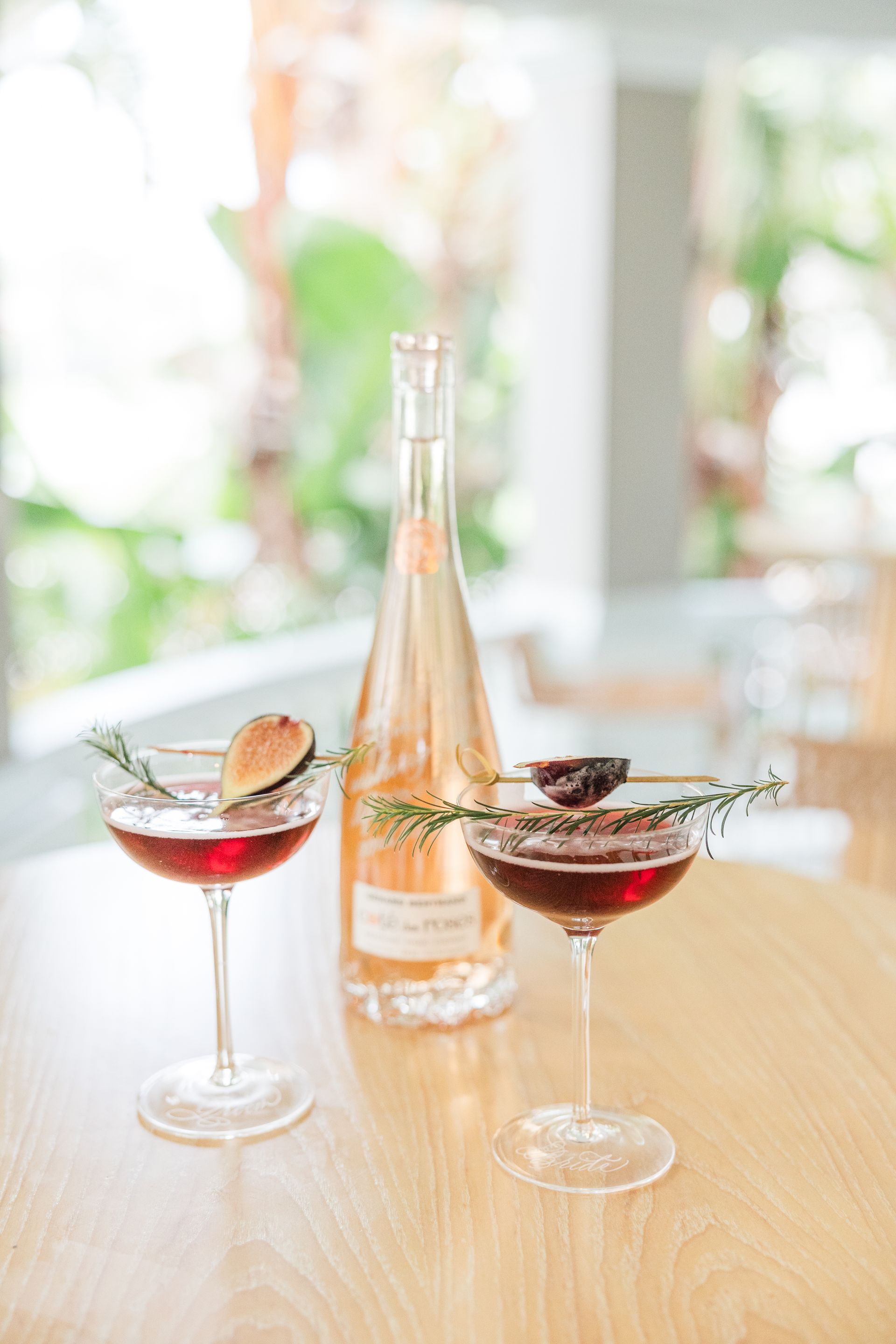 Two red cocktails with fig and rosemary garnish, a rose wine bottle on a wooden table.