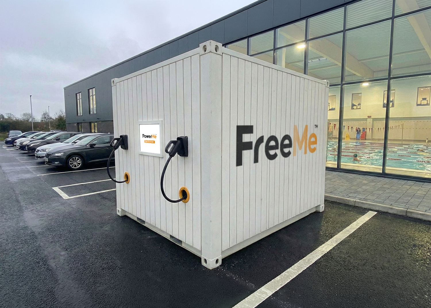 White ground work free FreeMe EV charging unit in a wet parking lot beside a glass-fronted building