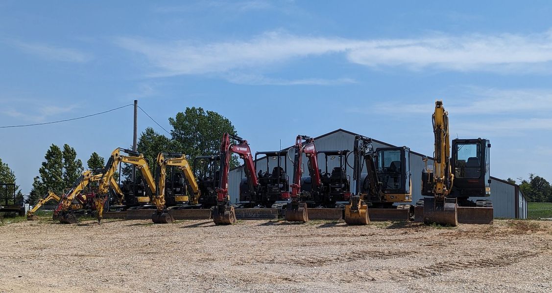 Row of excavators parked in front of a white building on a gravel lot, under a blue sky.