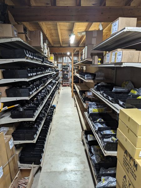 Warehouse aisle with shelves full of black items and boxes. Wooden ceiling and concrete floor.