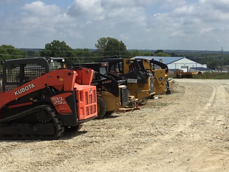 Several skid steer loaders parked on a gravel lot under a sunny sky.