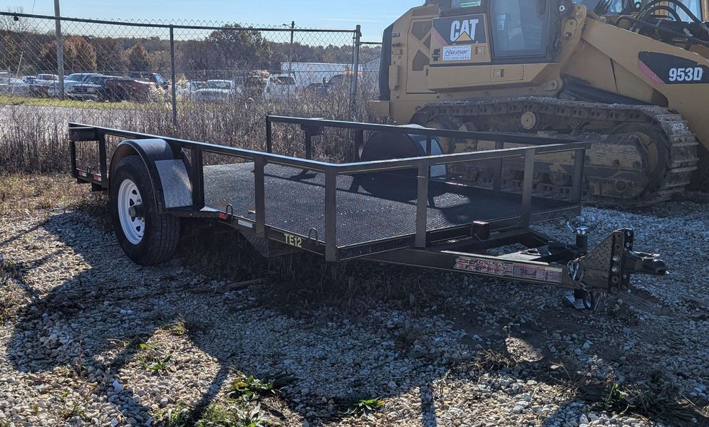 Black utility trailer with mesh sides parked outdoors, a yellow skid steer is in the background.