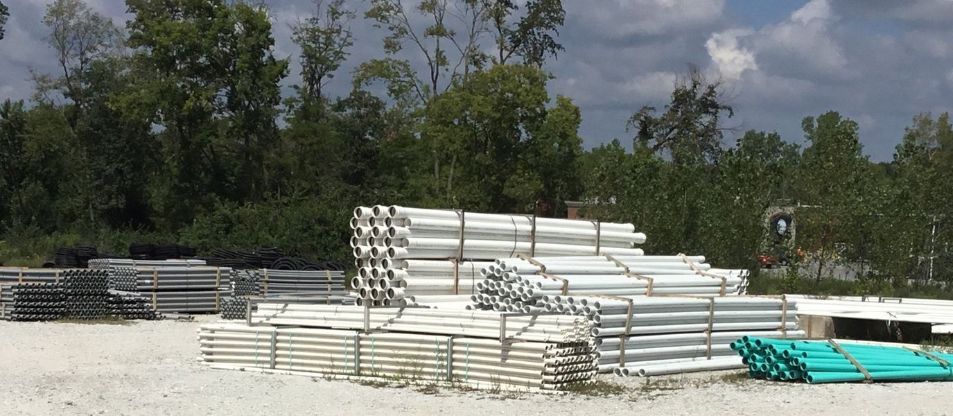 Piles of white and green pipes in an outdoor storage area, with trees in the background under a cloudy sky.