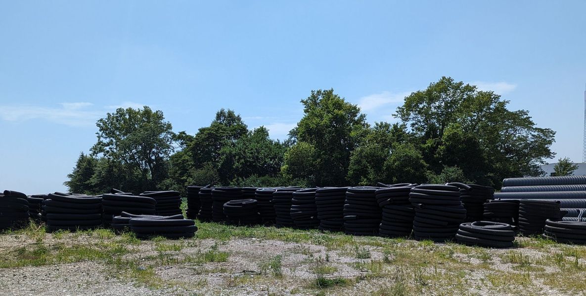 Stacks of dark, rectangular objects on a grassy lot with trees in the background under a blue sky.
