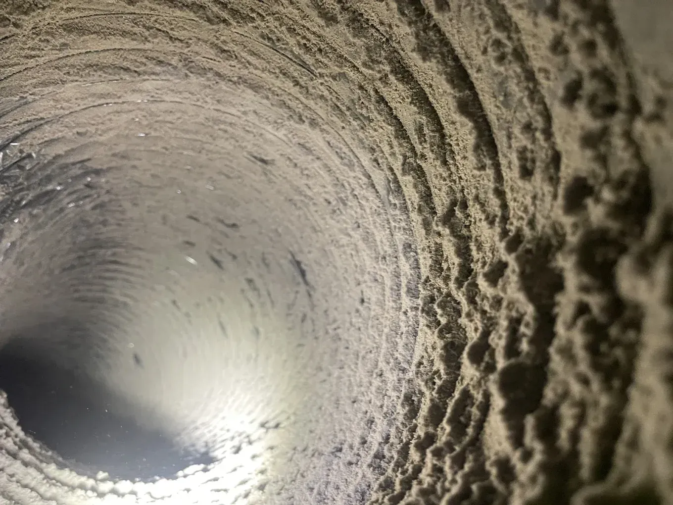Interior view of a dirty, cylindrical duct, coated with dust and debris. Light shines from end.