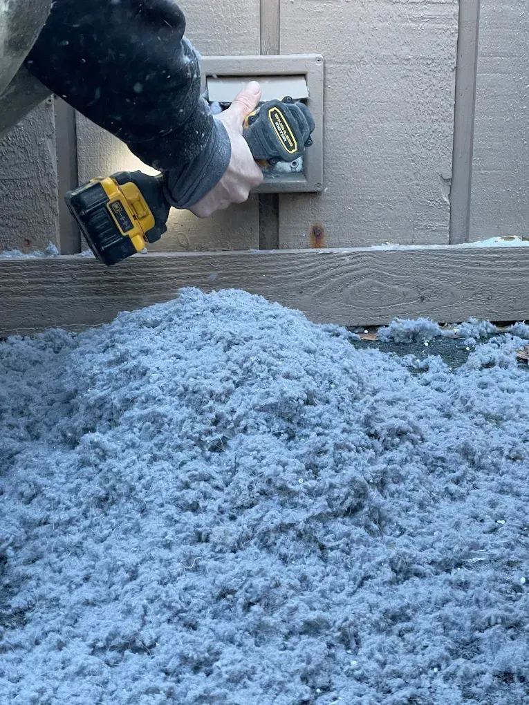 Person using a drill on a vent, with a pile of blue insulation below.