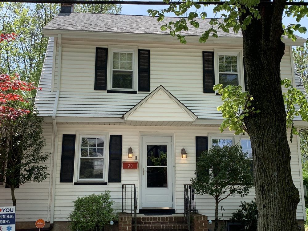 White two-story house with black shutters and a small front yard, surrounded by trees.
