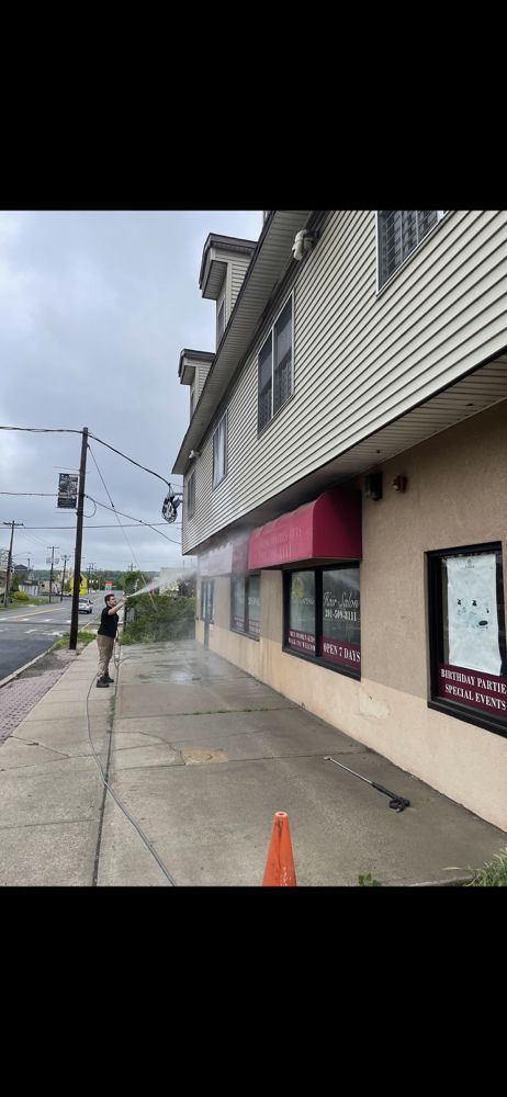 Person power washing the side of a building, sidewalk, and red awning. Orange cone in foreground. Overcast day.