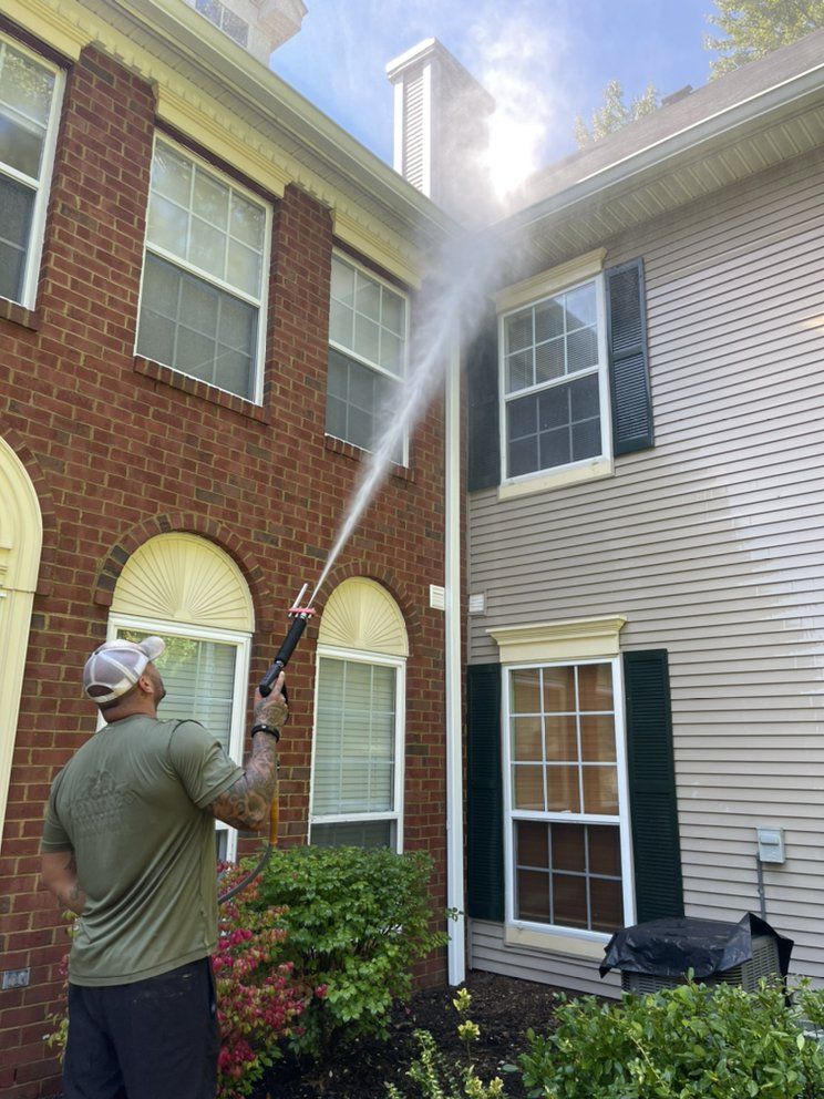 A person power washing the exterior of a two-story brick and siding house.