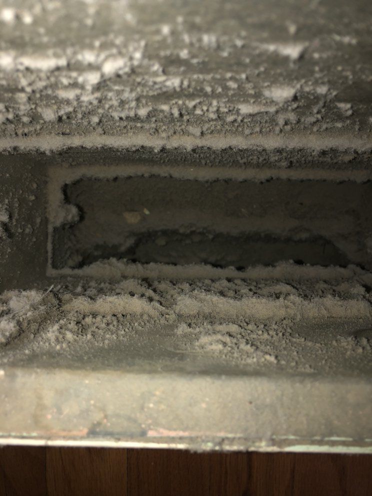 Close-up of a dusty air vent.  Gray dust and debris are caked inside the vent slats and surrounding edges.