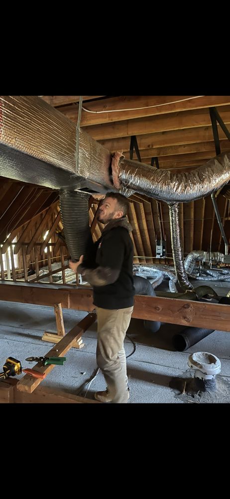 Man installing ductwork in an attic. He's holding a black pipe, surrounded by insulation and wooden beams.