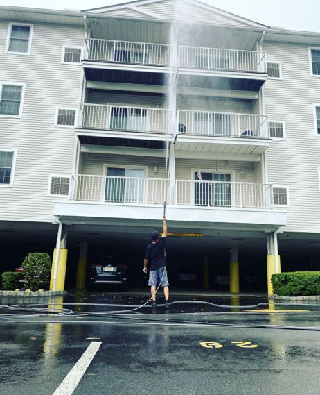Person pressure washing a multi-story apartment building exterior. Wet pavement reflects the building and sky.