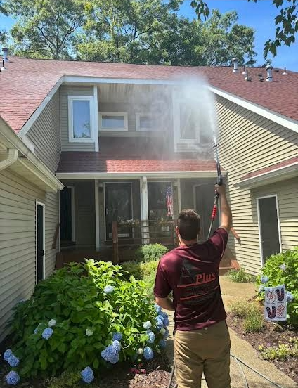 A person power washing the exterior of a two-story brick and siding house.