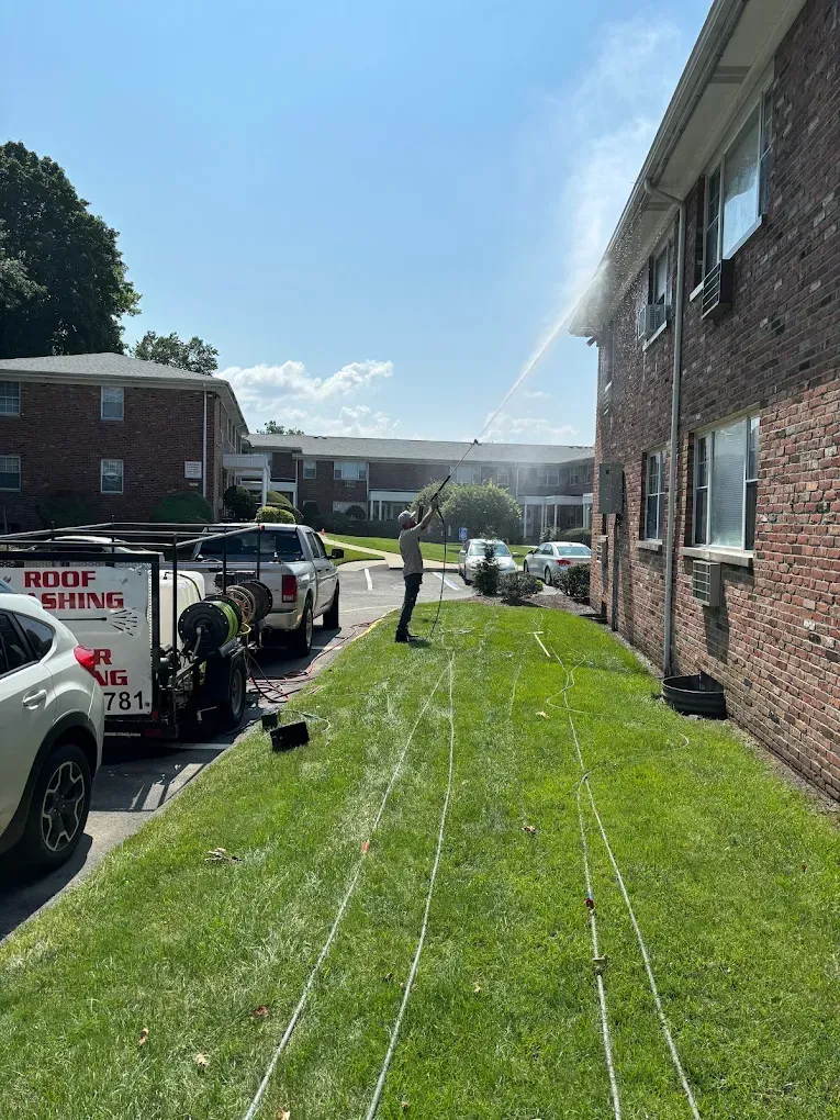 Man pressure washing a brick building on a sunny day. White truck and equipment are on the grass.