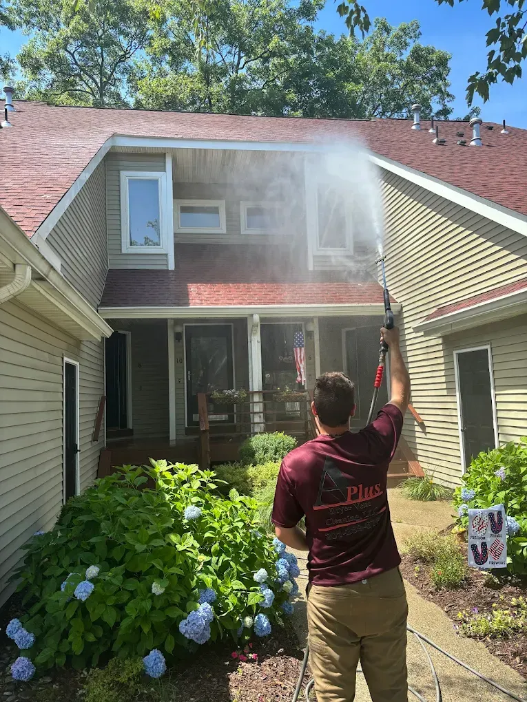 Person power washing a two-story beige house with a red roof, spraying water towards the siding.