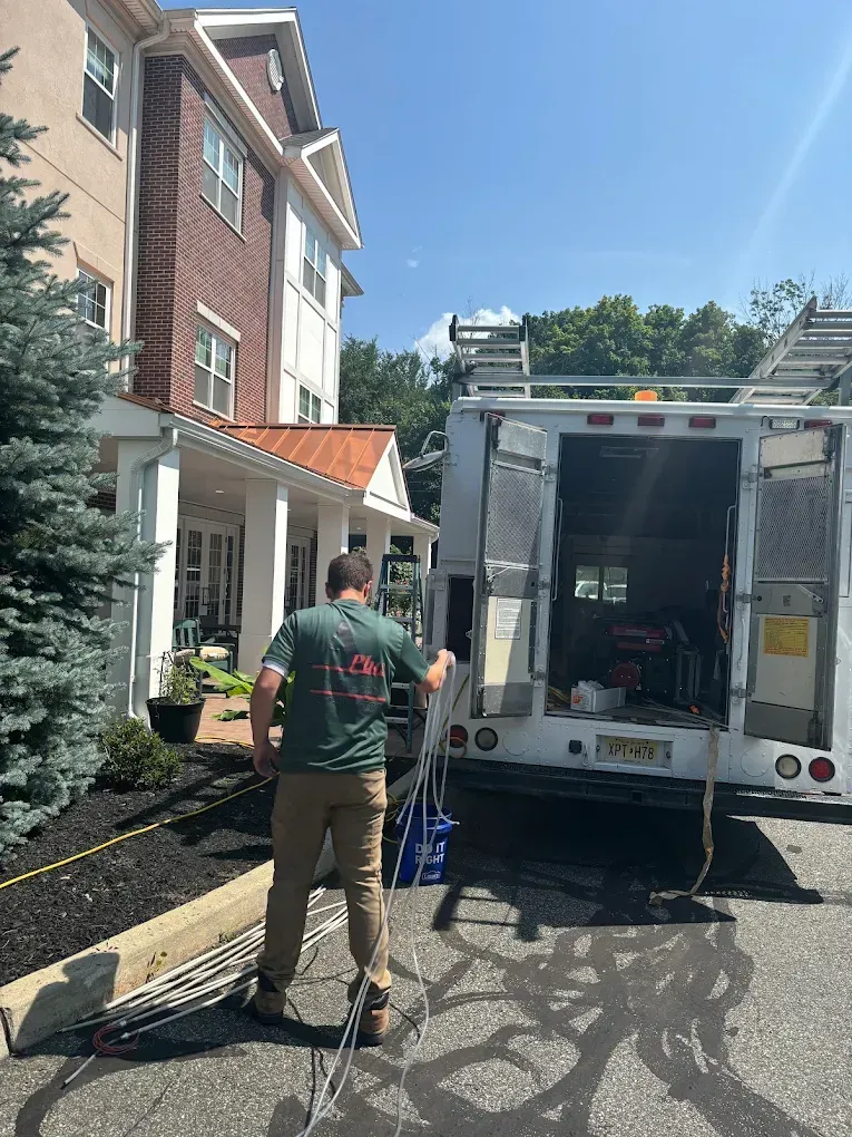 Man in green shirt near work truck, residential building in background. Sunny day.