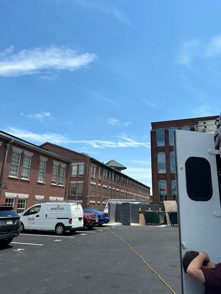 Brick buildings with parked vehicles under a blue sky, partially obscured by a white van and a person.