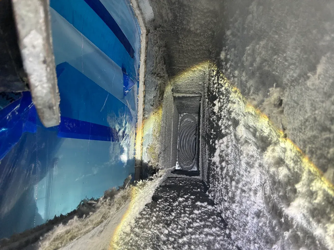 Narrow, dark duct interior with visible dust and debris. Blue taped wall visible on the left side, opening to a brighter area.