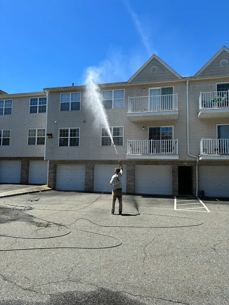 Person spraying water towards a building with a pressure washer on a sunny day.