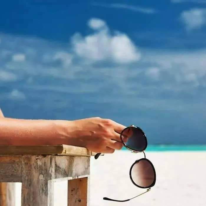 A person is holding a pair of sunglasses on the beach.