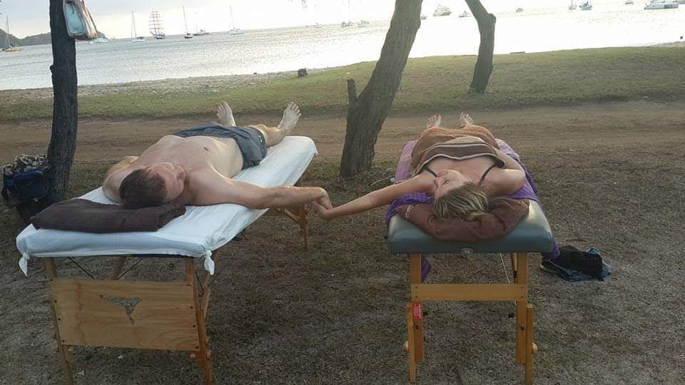 A man and a woman are laying on massage tables on the beach holding hands.
