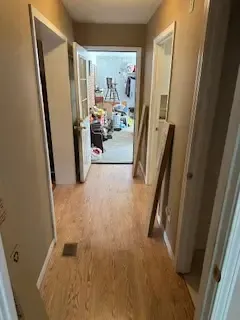 A hallway in a house with hardwood floors and a messy room in the background.