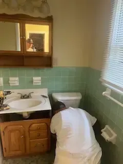 A man is kneeling in a bathroom next to a sink and toilet.