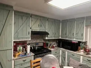 A kitchen with green cabinets , a stove , a microwave , and a chair.