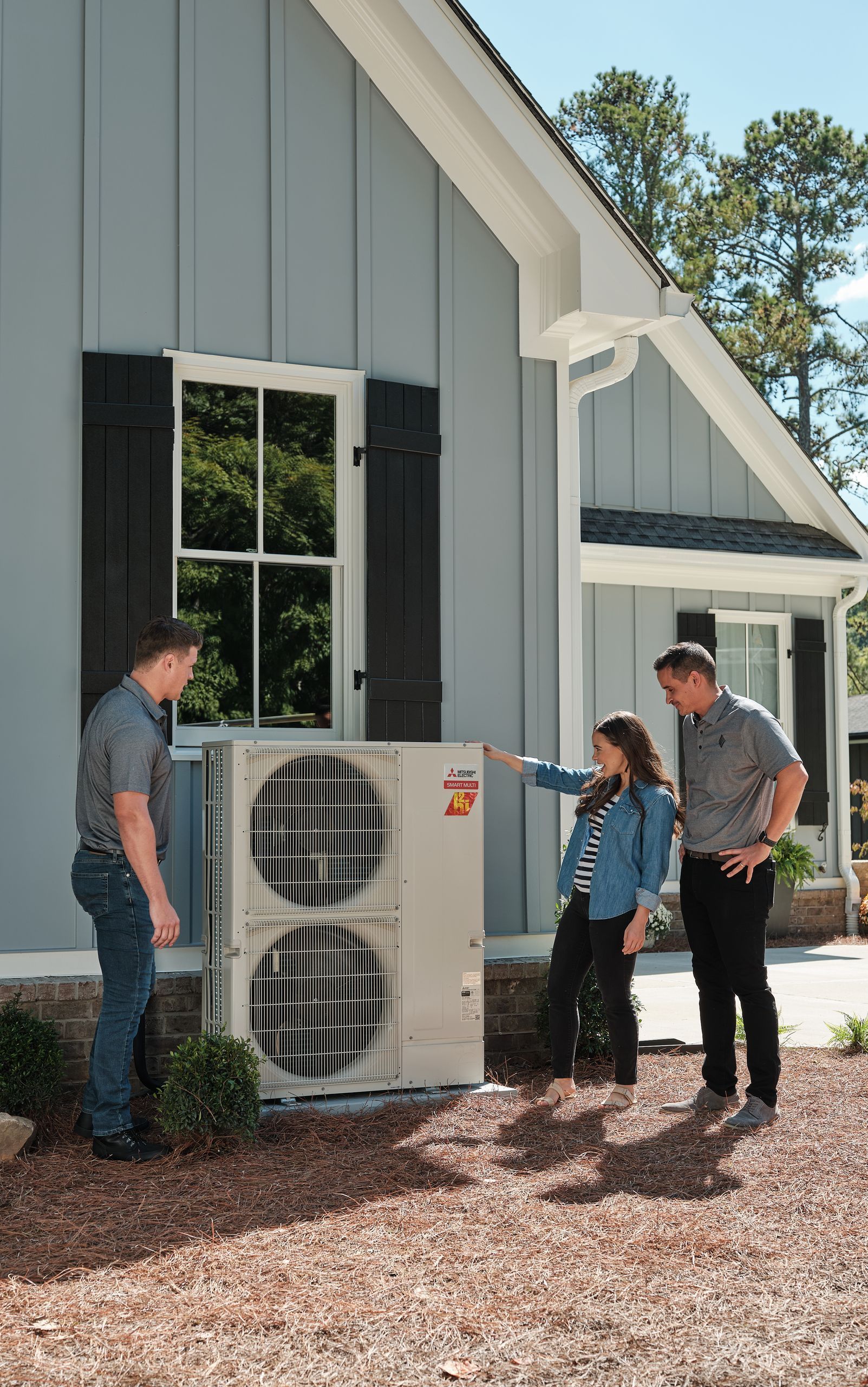 Three people examine an air conditioning unit outside a blue house. One points to the unit while the others look on.