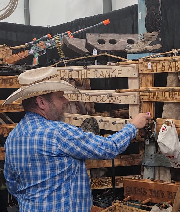 Man in a cowboy hat points at wooden art display with pallet shelves and toy guns.