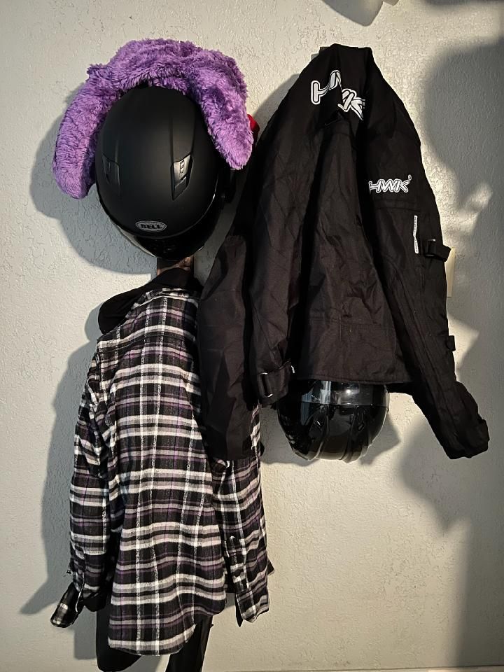 Black motorcycle helmets with purple fluffy accessory and jacket hanging on a wall.