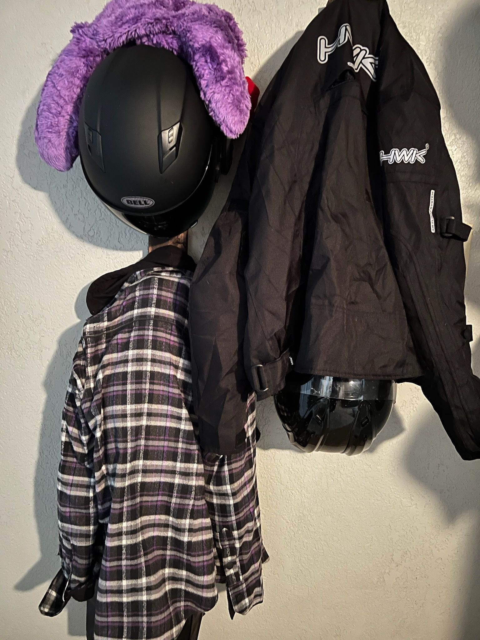 A motorcycle helmet with a purple cover and a black jacket hang on a wall, with a plaid shirt below.