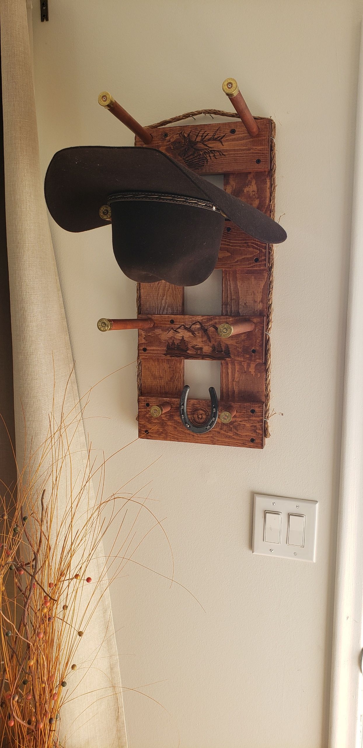 A cowboy hat hanging on a rustic wooden wall rack. The rack is mounted on a beige wall.