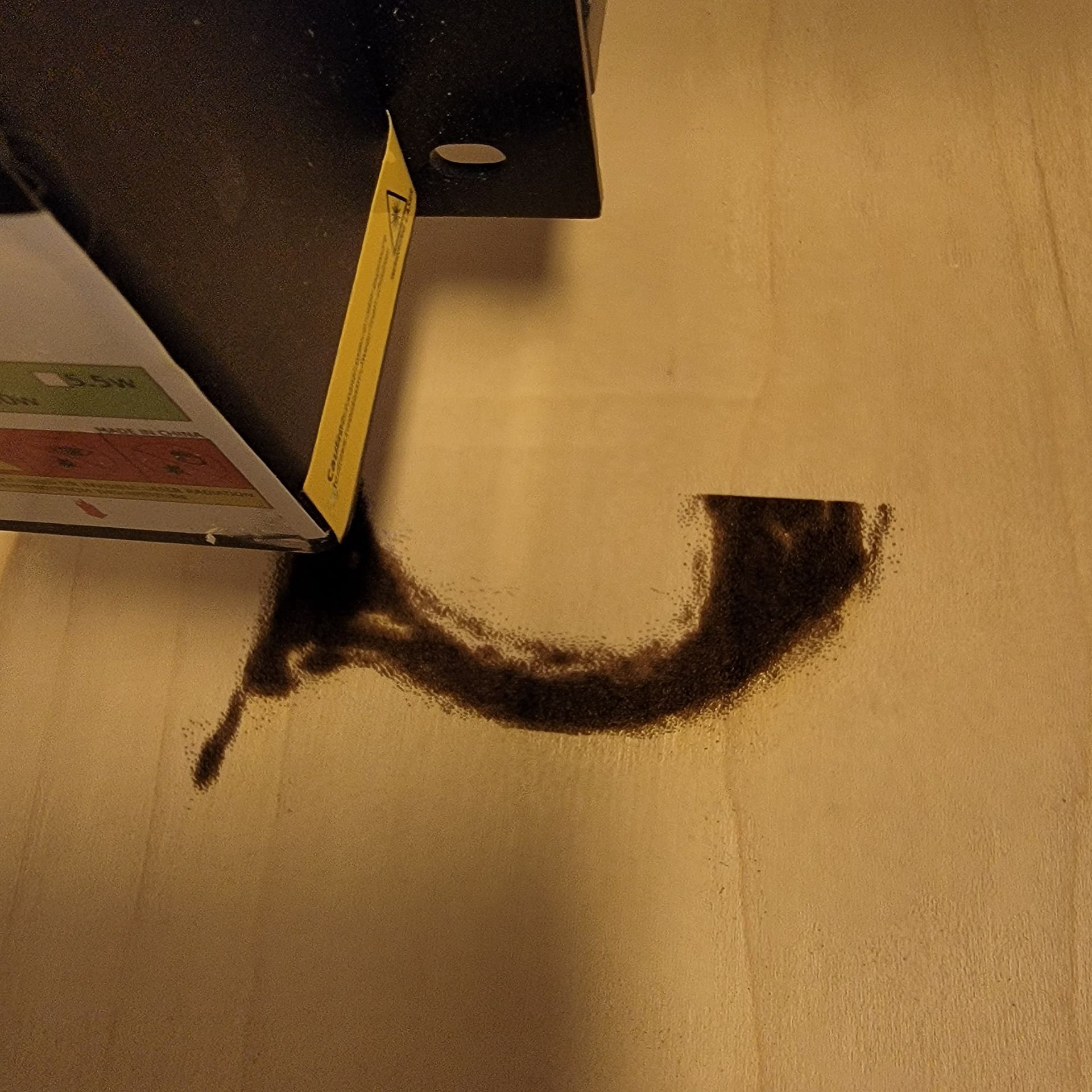 Laser cutter creating curved line of dark debris on wood. Black machine is in frame.