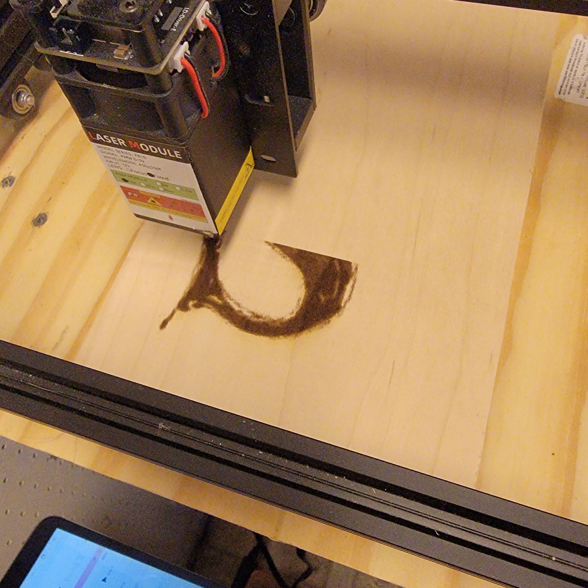 Laser engraver burning a curved shape into a light-colored wooden surface. The laser head is visible, along with a control screen.